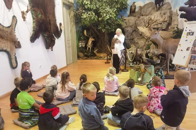 Un grup de copii stă pe perne pe podeaua unei încăperi cu diverse taxidermii de animale și elemente de decor naturale.