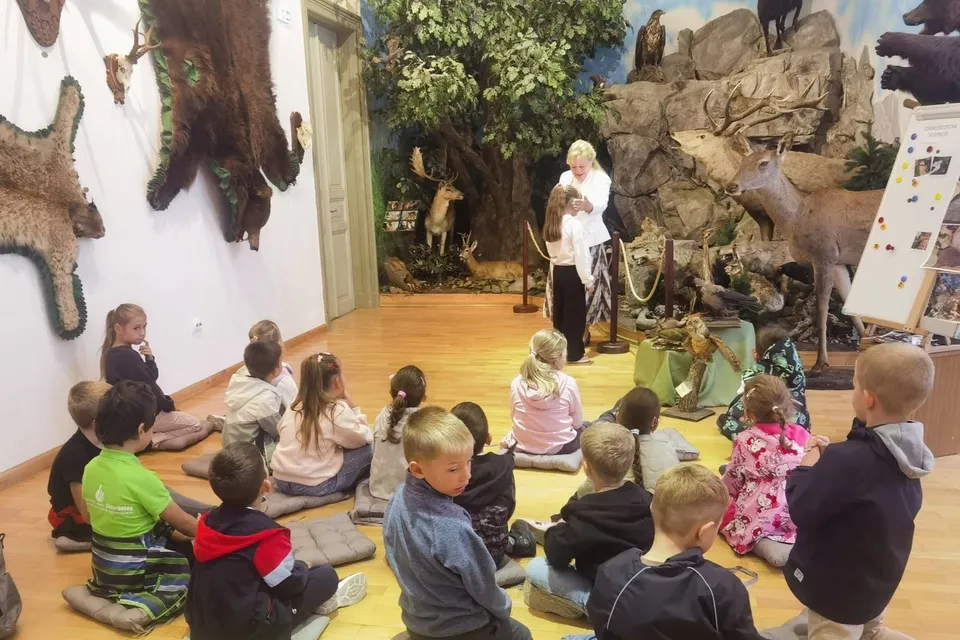 Un grup de copii stă pe perne pe podeaua unei încăperi cu diverse taxidermii de animale și elemente de decor naturale.