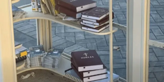 Mai multe cărți cu copertă maro și pagini albe sunt așezate pe rafturi albe de deasupra unei suprafețe pavate cu pietre gri.