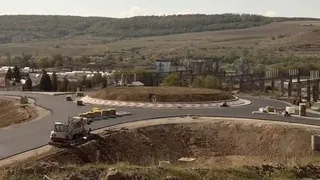 O mașină de compactat albă și gri lucrează pe un drum asfaltat lângă un rond de drum cu pământ și vegetație.