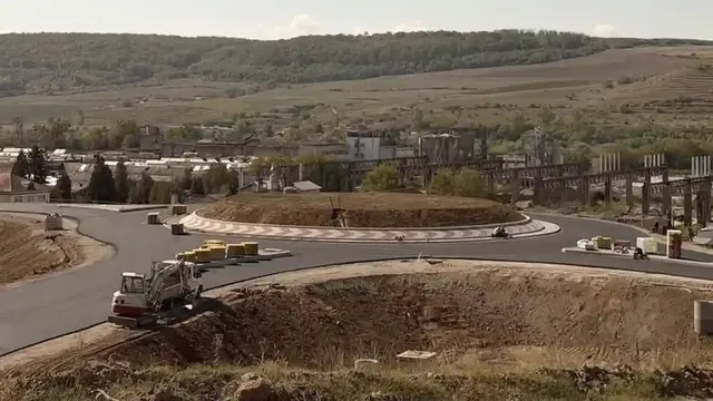O mașină de compactat albă și gri lucrează pe un drum asfaltat lângă un rond de drum cu pământ și vegetație.