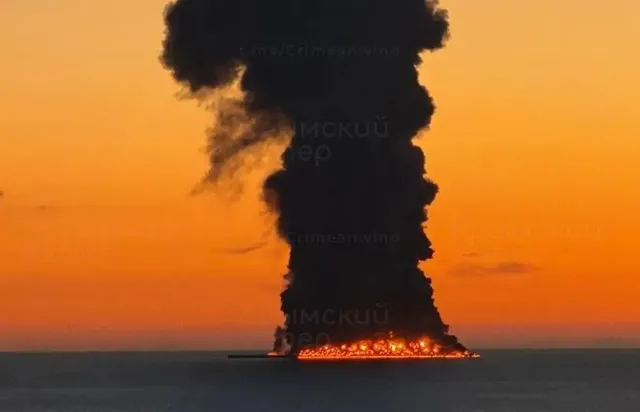 Un vas mare arde pe apă, emițând un nor dens de fum negru sub un cer portocaliu la apus.