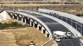 O autostradă curbată cu mai multe benzi de rulare este traversată de un camion cu betonier și un vehicul alb sub un cer senin.