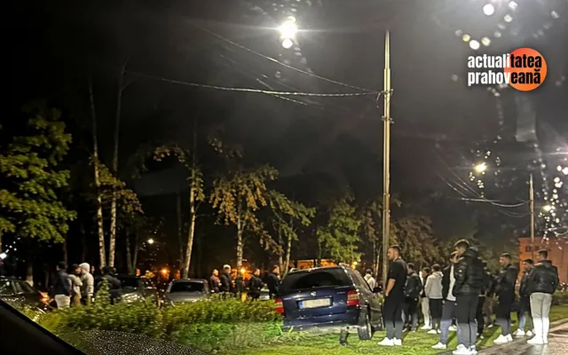 O mașină albastră este parcată pe un spațiu verde cu arbori în jurul unei străzi illuminate de lămpi la noapte, înconjurate de mai mulți oameni.