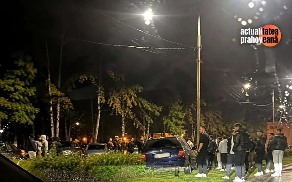 O mașină albastră este parcată pe un spațiu verde cu arbori în jurul unei străzi illuminate de lămpi la noapte, înconjurate de mai mulți oameni.