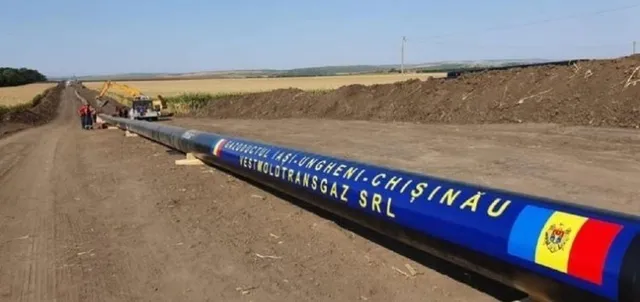 O conductă albastră cu inscripții albe și un steag colorat este întinsă pe un teren de pământ lângă un utilaj de excavare.