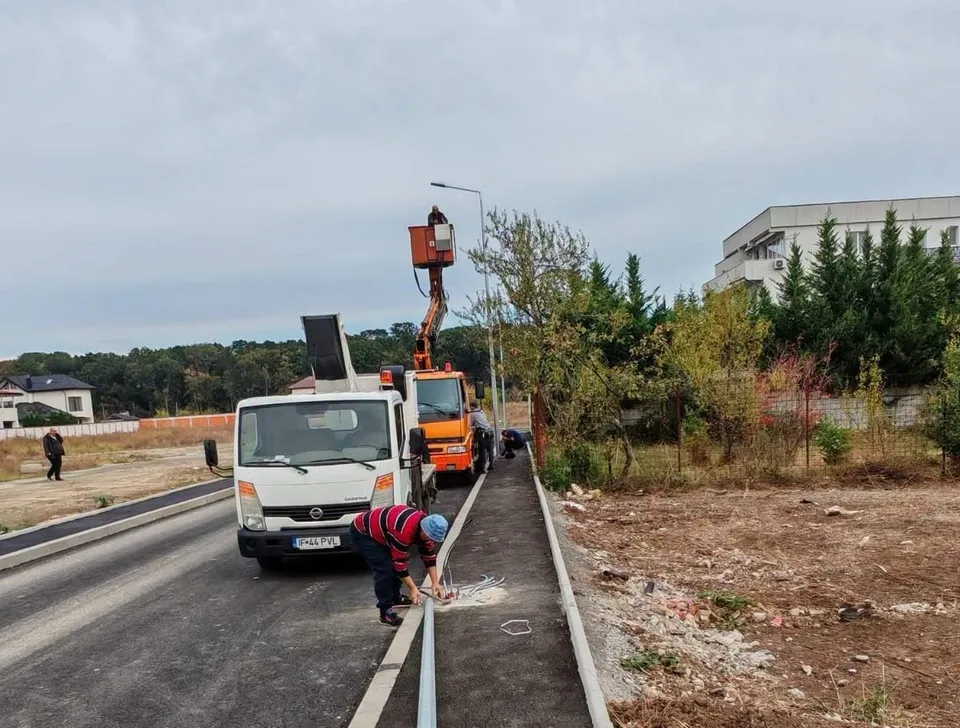 Un lucrător cu vestă roșie și albastră este aplecat lângă bordură pe un drum asfaltat, lângă un camion alb și un vehicul utilitar portocaliu.