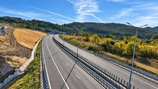 O autostradă cu două benzi de fiecare sens trece printr-un peisaj cu dealuri împădurite sub un cer albastru cu câteva nori subțiri.
