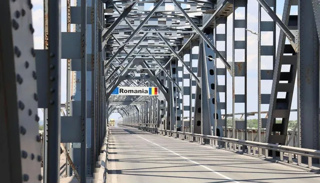 O perspectivă de la nivelul drumului arată o structură metalică de pod cu grinzi și stâlpi gri sub un cer senin.