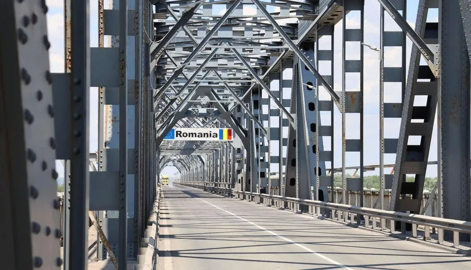 O perspectivă de la nivelul drumului arată o structură metalică de pod cu grinzi și stâlpi gri sub un cer senin.
