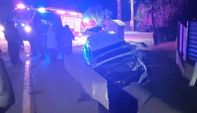 O mașină albă este lovită de un gard de beton sub lumini albastre de serviciu de urgență, în timp ce persoane stau lângă vehicul.